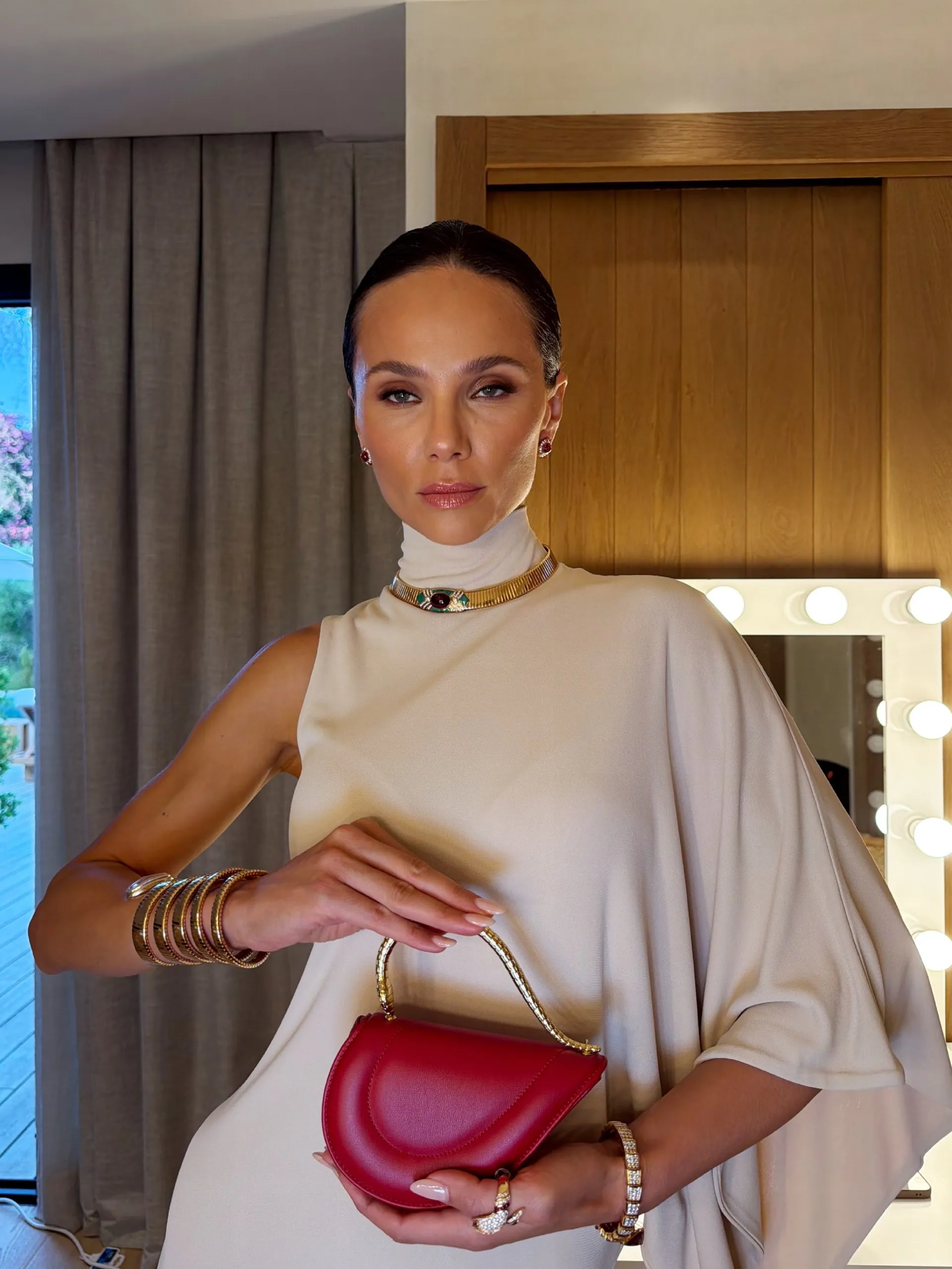 Модель в кремовом платье с красной сумкой Bvlgari и золотыми браслетами